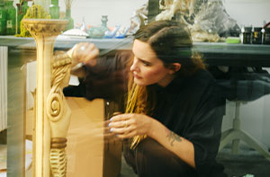 Une artiste, Bianca Bondi, agenouillée dans un atelier, sculpte ou restaure une colonne décorative ornée d’un serpent doré, entourée d’objets artistiques, de fleurs séchées et d’éléments naturels.