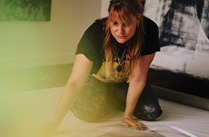 Eva Nielsen, a contemporary artist, kneeling in her studio, focused on a floor-based artwork, surrounded by abstract paintings, with a soft light filter in the foreground.